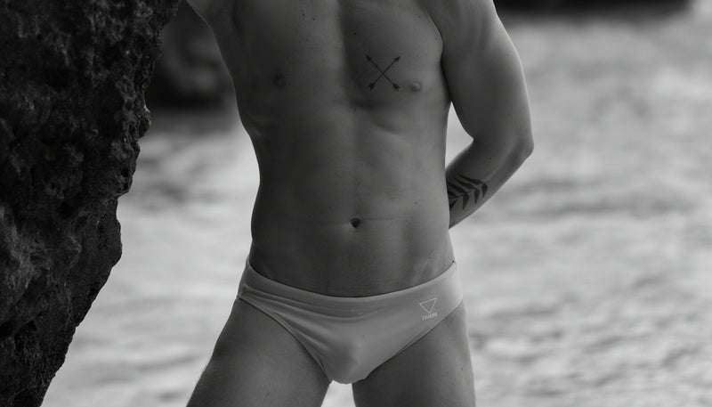 Man wearing blue swim trunks standing by a rocky shore with water in the background